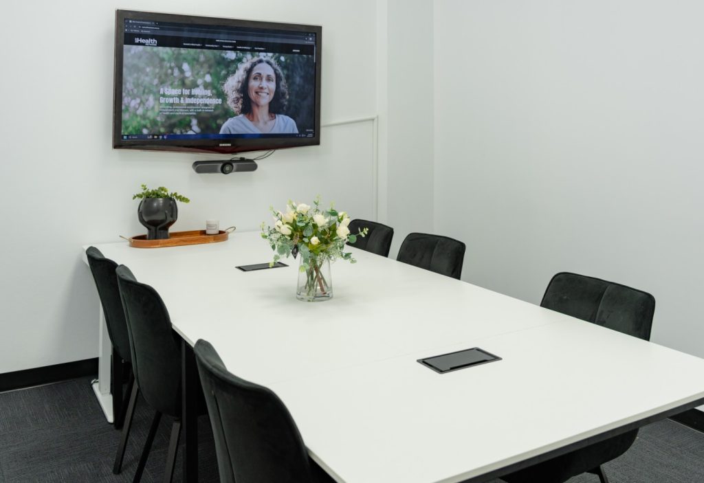 Group room at My Health Network featuring a white table, black chairs, a TV screen for presentations, and a floral centerpiece, suitable for workshops, therapy sessions, and community events.