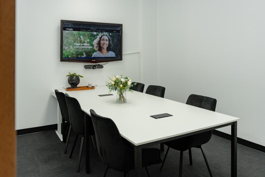 Consulting room at My Health Network featuring a white table, black chairs, a floral centerpiece, and a TV displaying health services information.