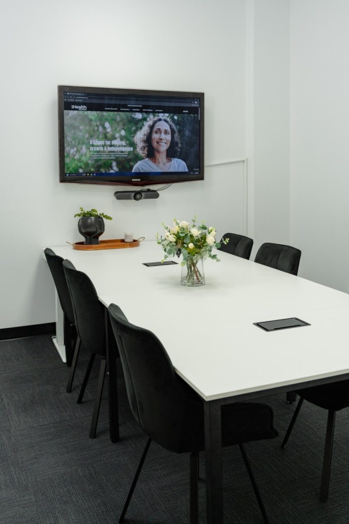 Group room with a large white table, black chairs, and a TV displaying My Health Network website, featuring a floral centerpiece, suitable for workshops and group activities.