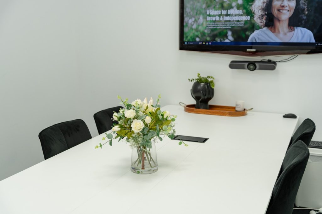 Group room featuring a white table with a floral arrangement, black chairs, and a TV screen displaying "A Space for Healing, Growth & Independence," equipped for presentations and workshops.