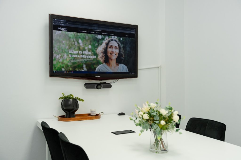 Group room at My Health Network featuring a TV screen for presentations, a video conference camera, a modern table, black chairs, and a floral centerpiece, emphasizing a welcoming environment for workshops and group activities.