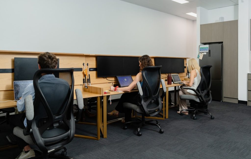 Three health professionals working at hot desks in a modern office space, featuring ergonomic chairs, computers, and collaborative work environment, emphasizing flexibility and community for allied health practitioners.