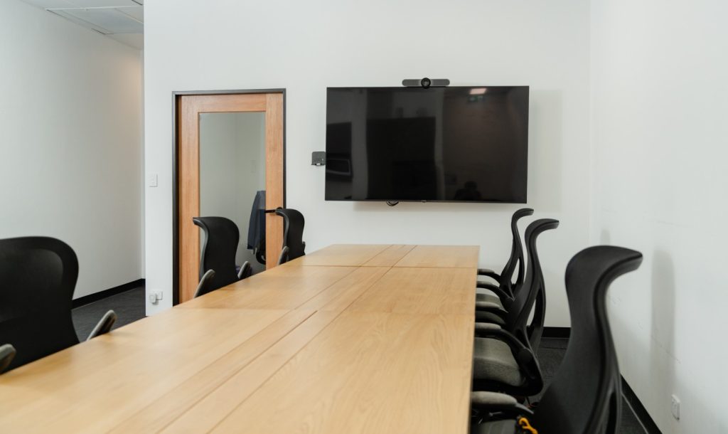 Meeting room with a long wooden table, ergonomic black chairs, and a large wall-mounted screen, suitable for collaborative work among health professionals at My Health Network.