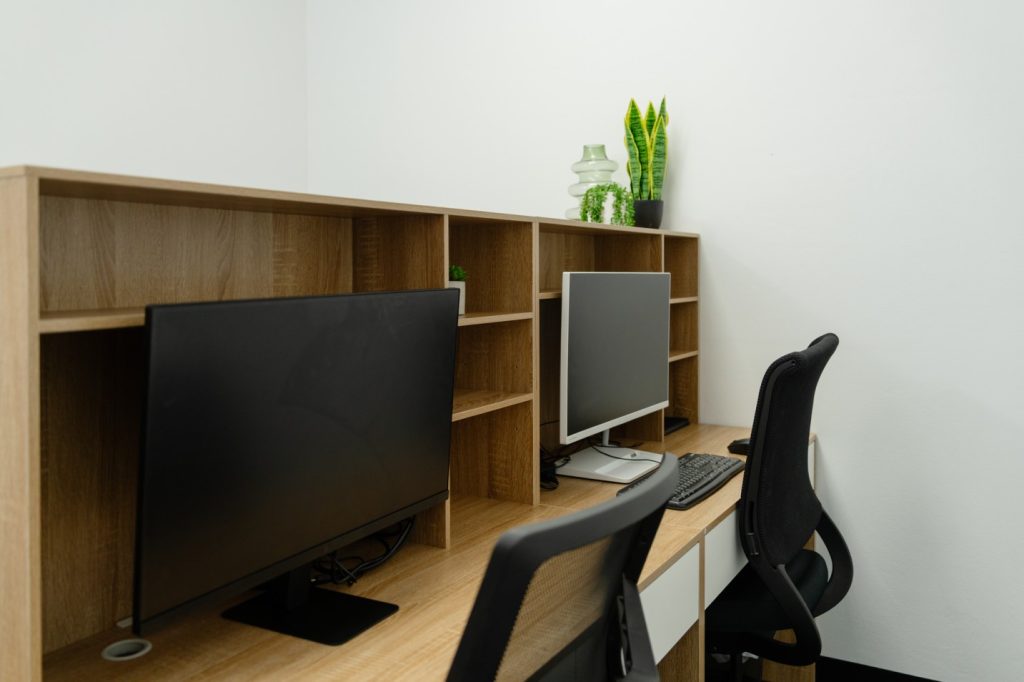 Hot desk workspace featuring two computers, ergonomic chairs, wooden shelving, and decorative plants, ideal for healthcare professionals seeking flexible work solutions.