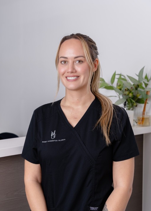 Registered Nurse Michelle Boyce in a black medical uniform, smiling confidently in a cosmetic clinic setting, with plants and treatment tools in the background, representing personalized aesthetic care and expertise in cosmetic injectables.