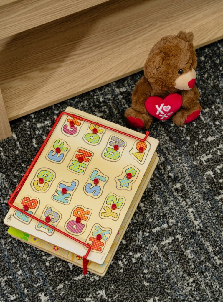 Colorful children's learning book with letters and shapes, alongside a plush teddy bear with a heart, on a textured carpet, reflecting a nurturing environment for therapy and child development.
