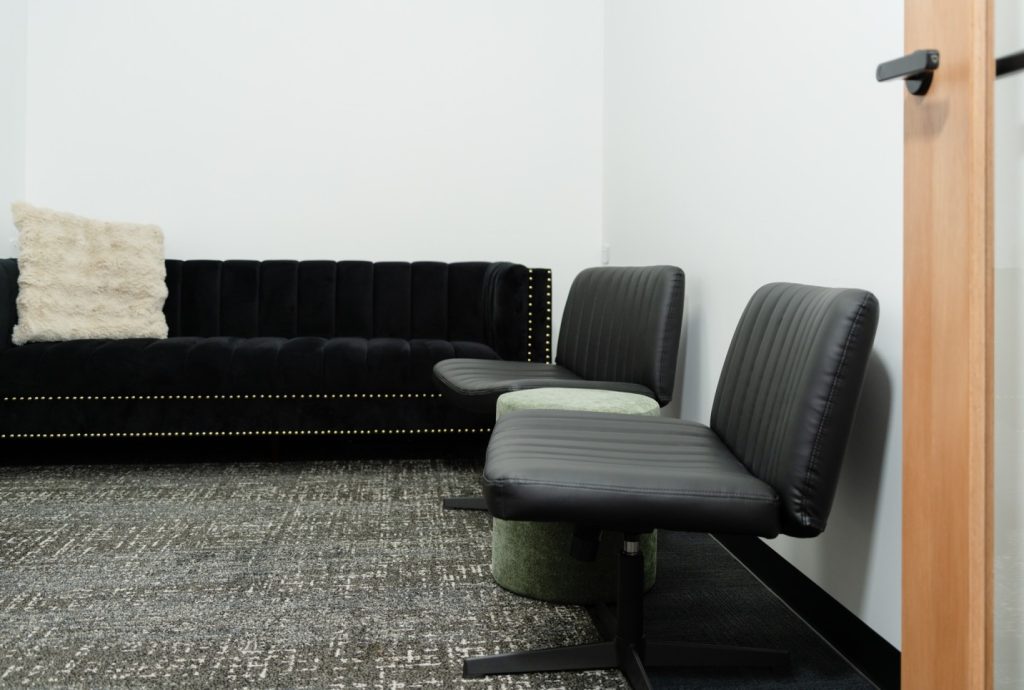 Therapy room interior featuring a black velvet couch with decorative pillow, two modern black chairs, and a green ottoman, emphasizing privacy and comfort for health practitioners and clients.
